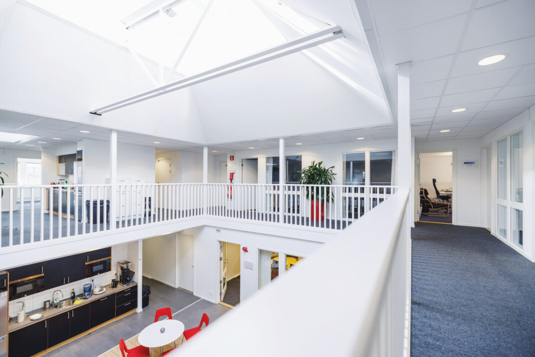 Modern kontorsinredning med takfönster, öppet kök, runt bord med röda stolar och en korridor med kontor och växter.