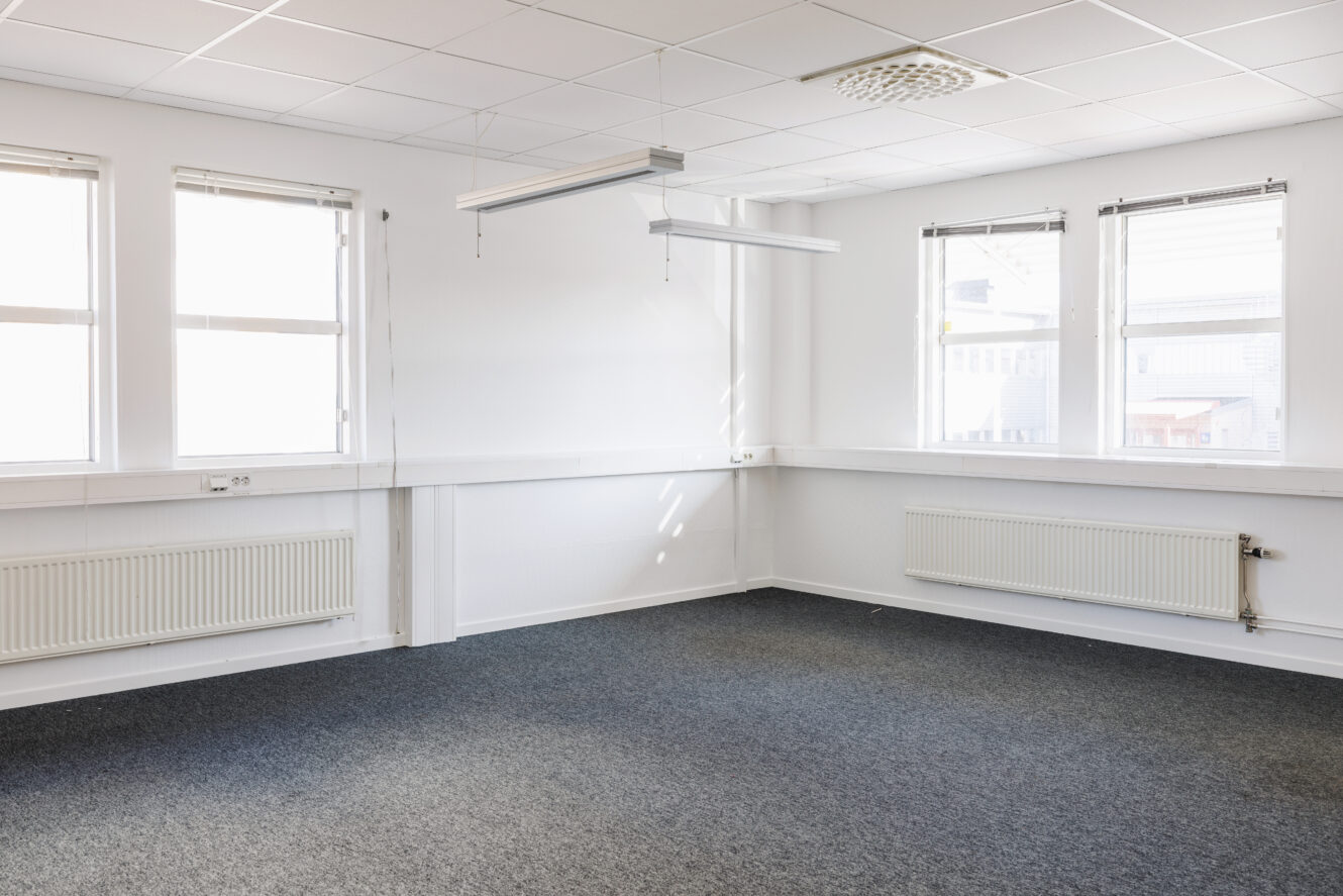 Tomt kontorsrum med vita väggar, tre fönster, två radiatorer, upphängda lysrör och grå heltäckningsmatta.