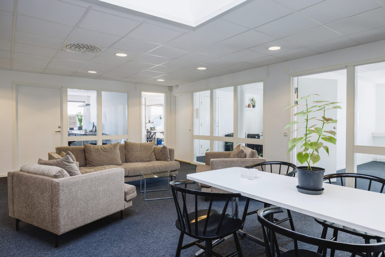 Modern kontorslounge med gråa soffor, ett vitt bord omgivet av svarta stolar, en krukväxt och kontor med glasväggar i bakgrunden.