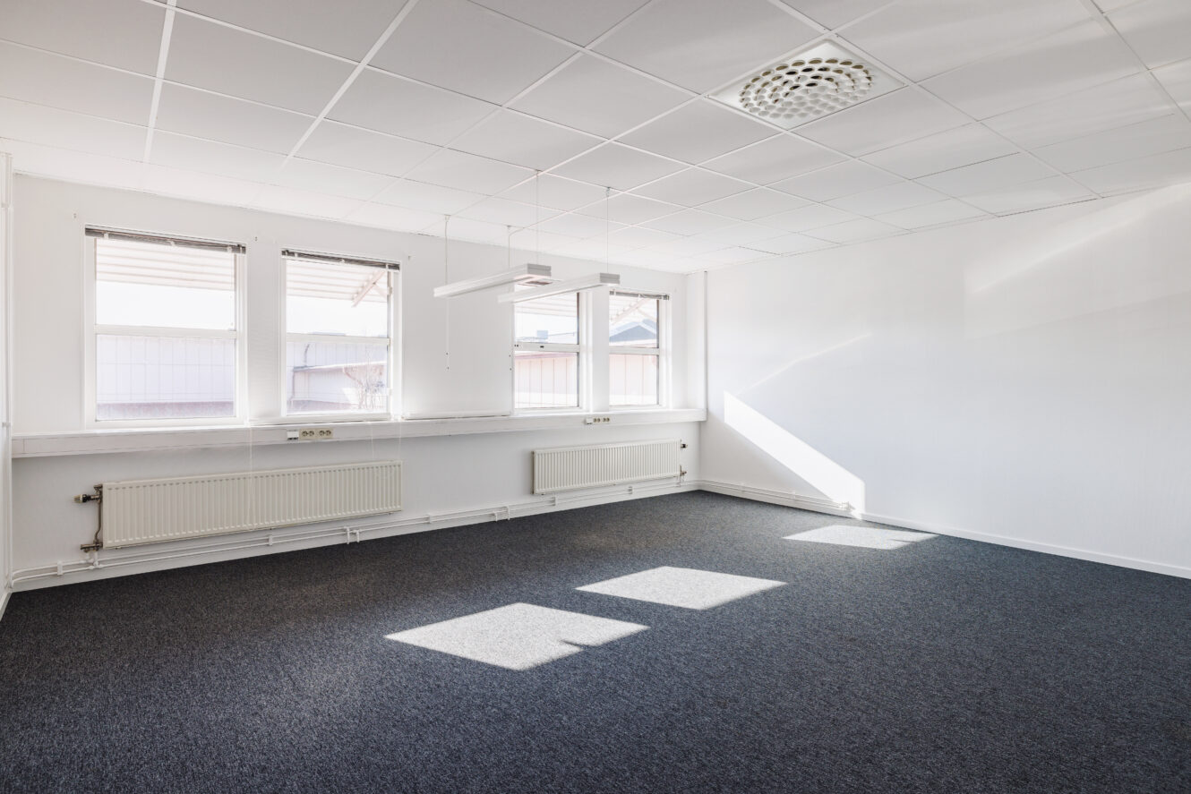 Tomt kontorsrum med tre stora fönster, vita väggar, heltäckningsmatta, taklampor och radiatorer under fönstren. Solljuset kastar skuggor på golvet.