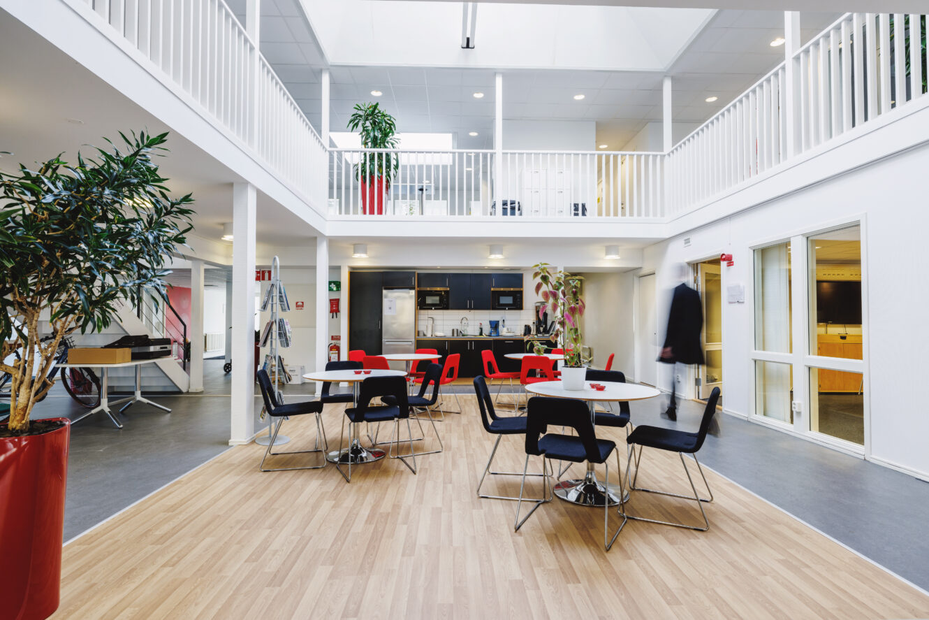 Modernt kontorskök och matplats med svarta och röda stolar, bord, växter och en mezzanin-nivå under ett takfönster.