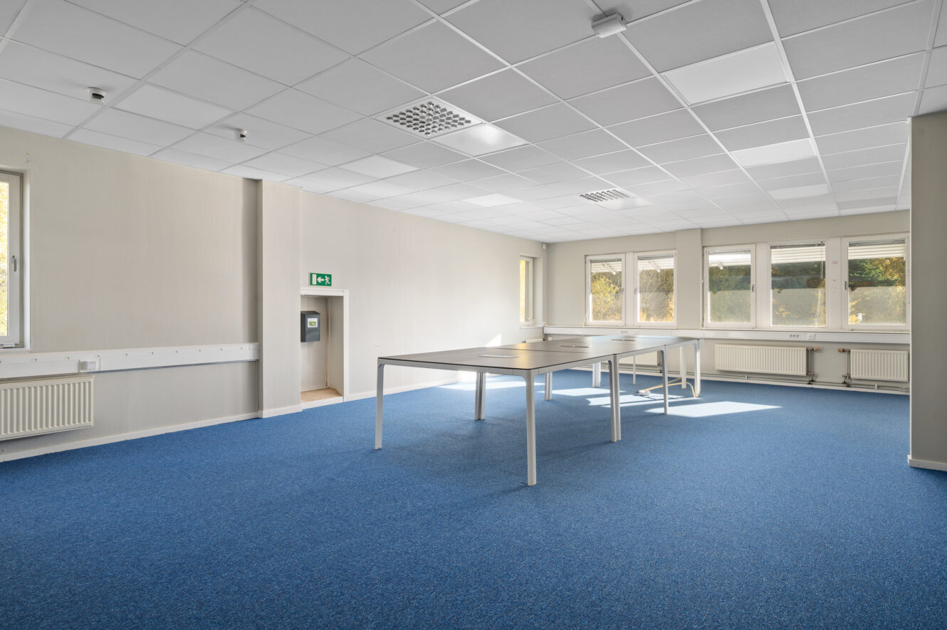 Ett rymligt, tomt kontorsrum med blå heltäckningsmatta, vita väggar, stora fönster och flera vita bord uppställda i mitten.