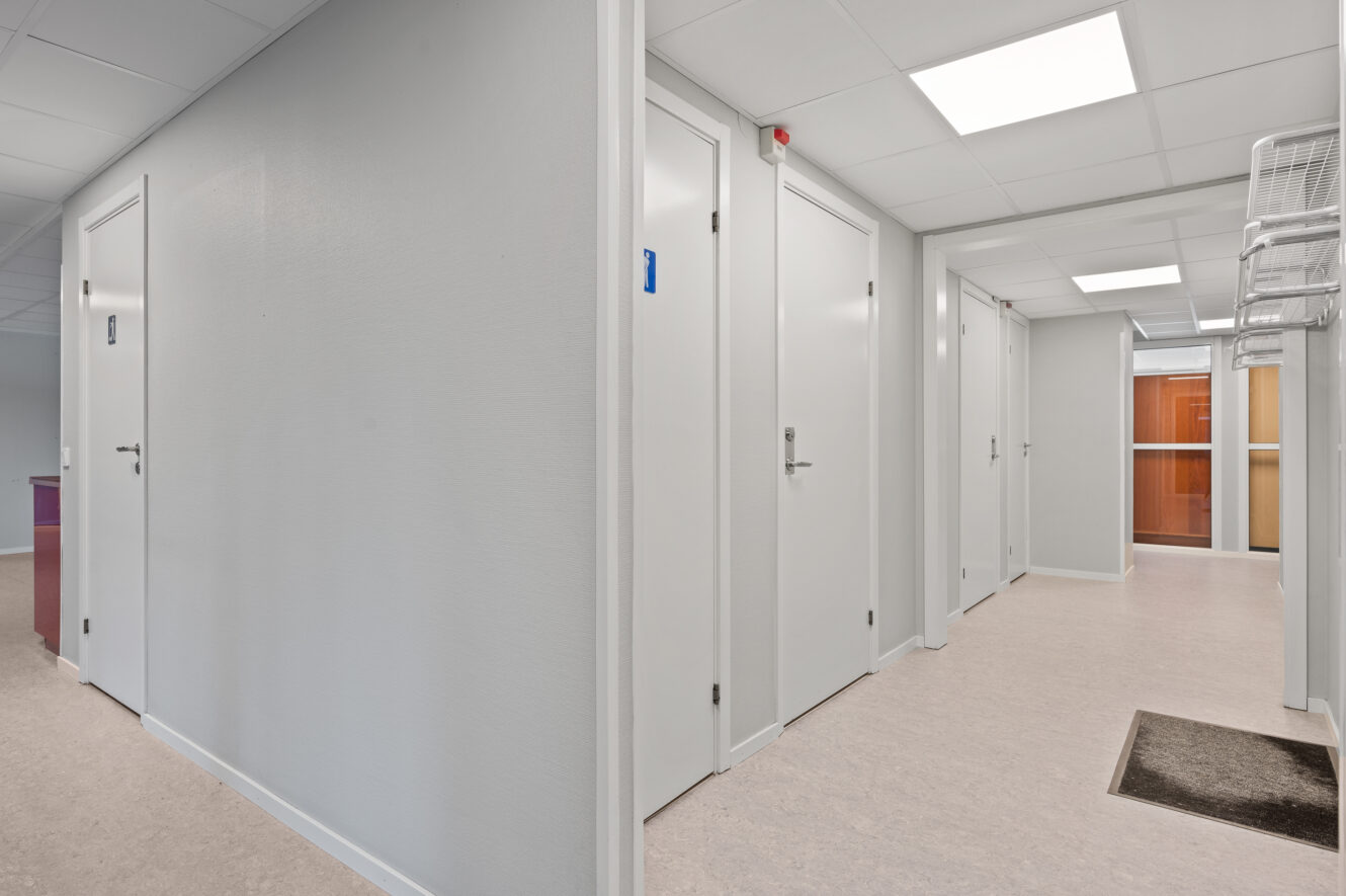 En ren, modern hall i en kontors- eller vårdbyggnad med stängda vita dörrar, neutrala väggar och taklampor.