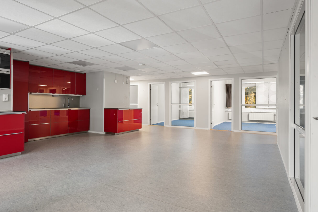 Modernt tomt rum med röda köksskåp, grått golv, vita väggar och ett nedfällt tak. Stora fönster och glasdörrar ger naturligt ljus och utomhusutsikt.