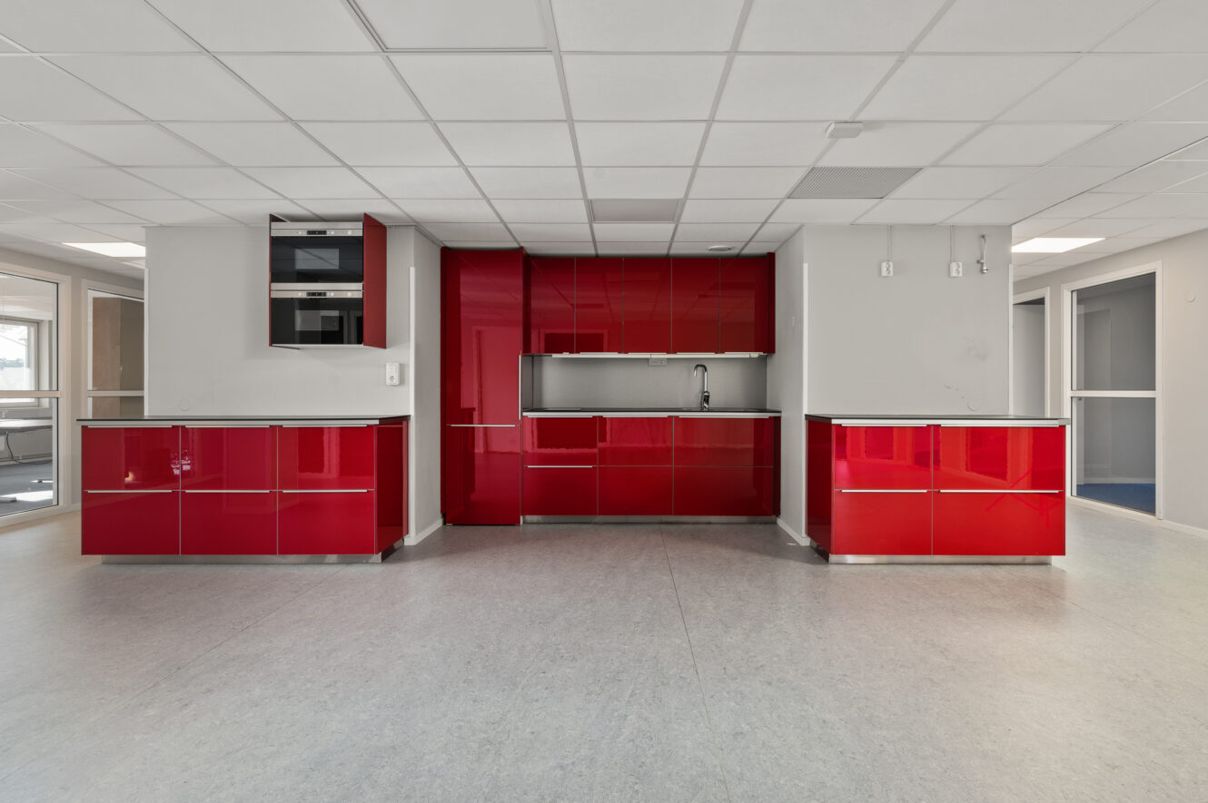 Modernt kök med blanka röda skåp, grå bänkskivor, en inbyggd diskho och neutrala väggar; öppen planlösning med gott om naturligt ljus från fönster.