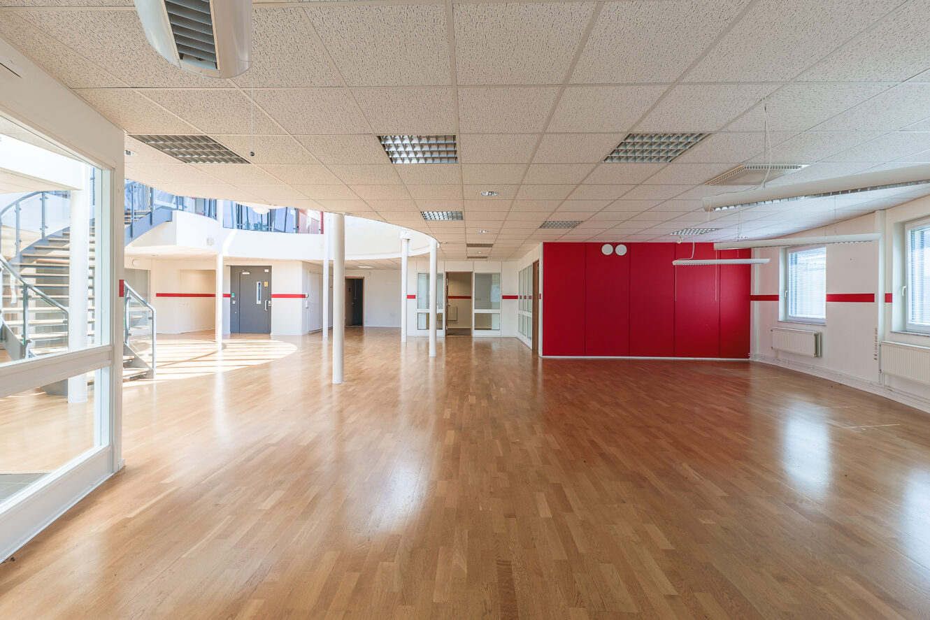 Rymligt, tomt rum med trägolv, röd accentvägg, stora fönster och en spiraltrappa i bakgrunden under ett gallertak med lysrör.