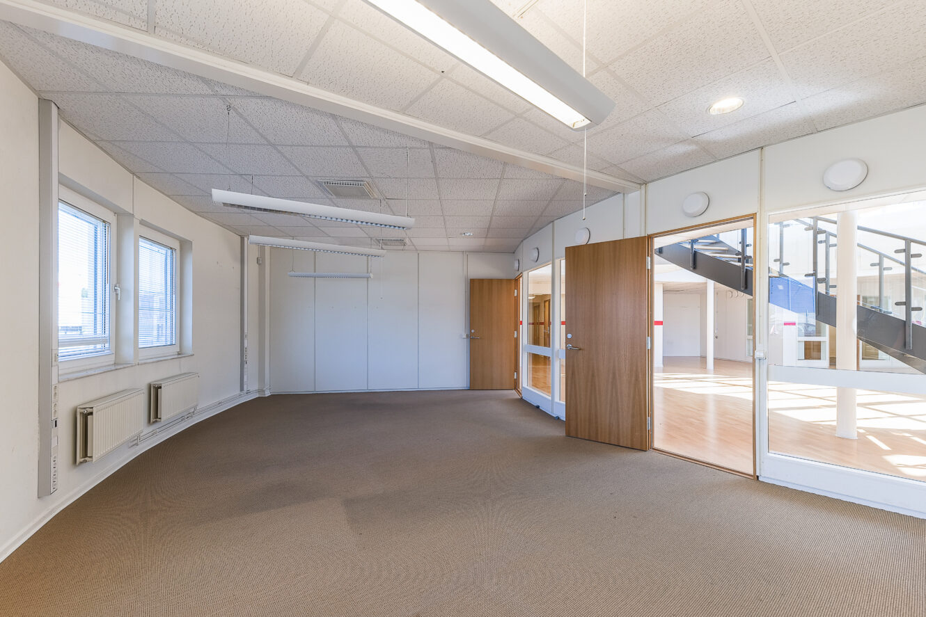 Tomt kontorsutrymme med beige matta, vita väggar, lysrörsbelysning och stora fönster; glasdörrar leder till ett angränsande område med trappor och trägolv.
