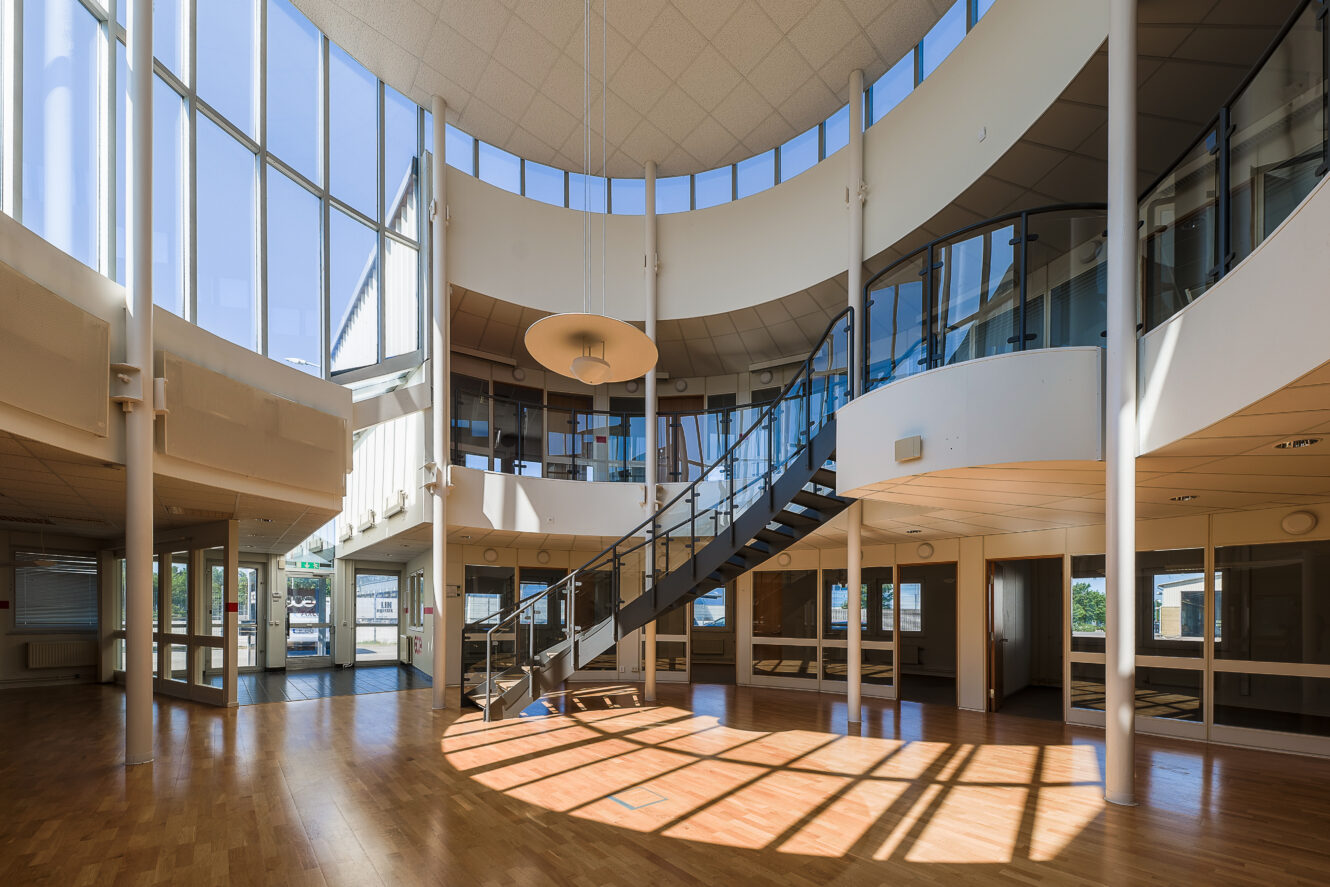 Rymlig modern interiör med stora fönster, trägolv, central trappa och glasväggar på övre plan; solljuset flödar in genom fönstren.