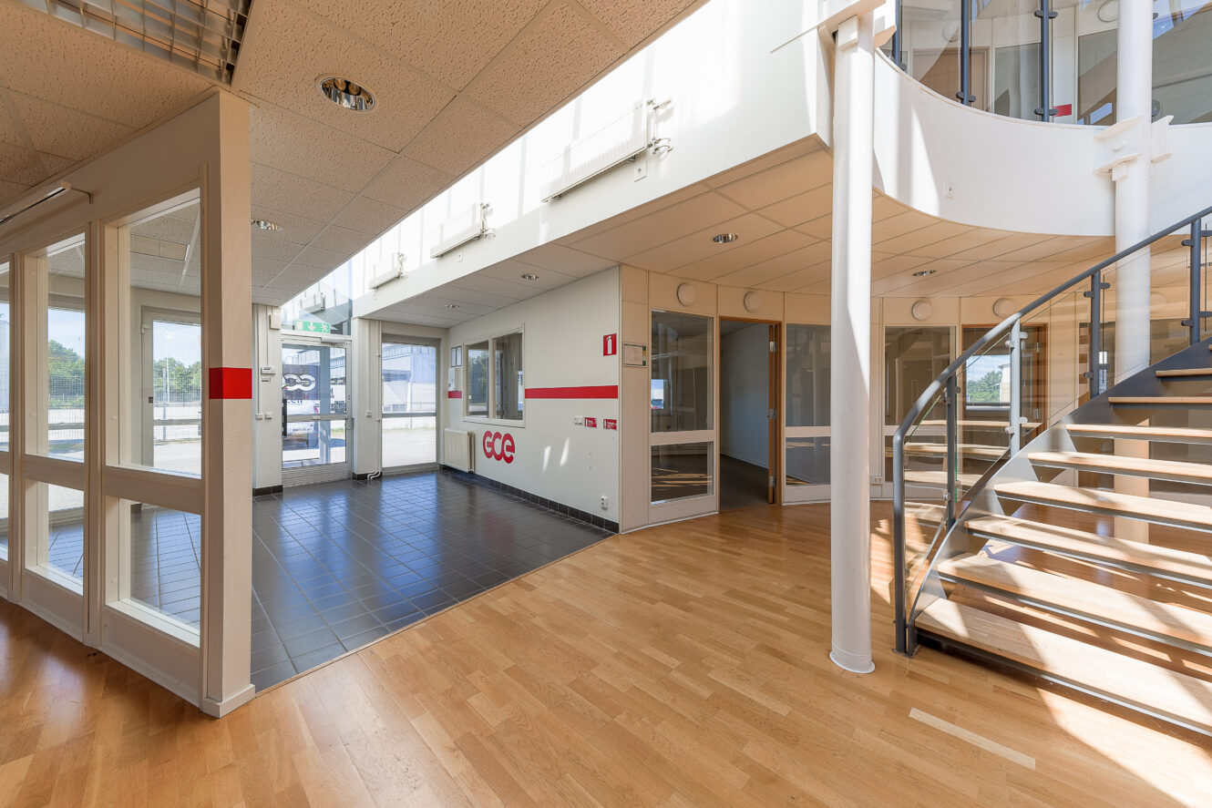 En modern kontorsinredning med trä- och klinkergolv, glasväggar, en spiraltrappa och rikligt med naturligt ljus från stora fönster.