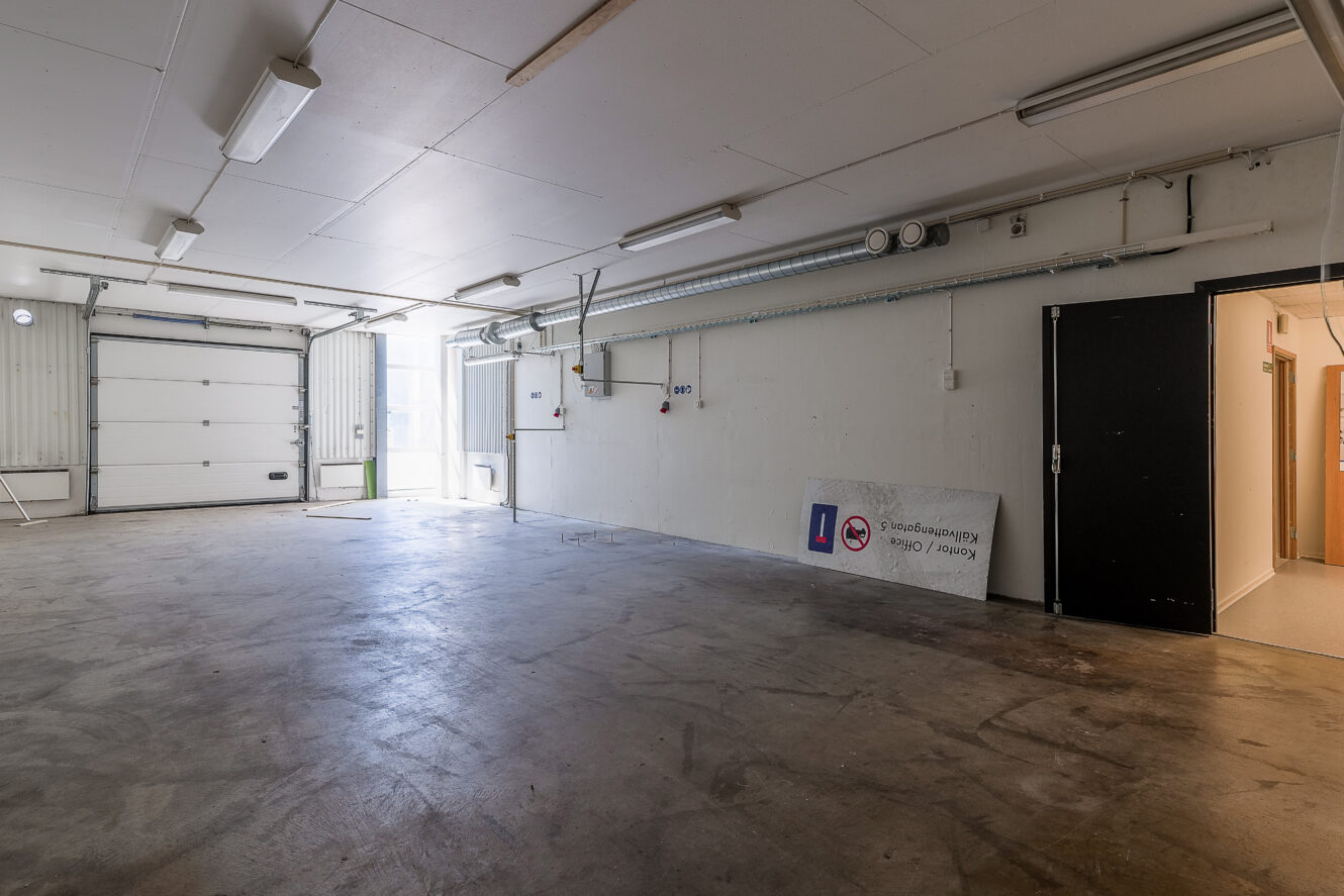 Tomt garage med betonggolv, vita väggar, takbelysning, delvis öppen garageport och en öppen sidodörr; en stor skylt vilar mot väggen till höger.