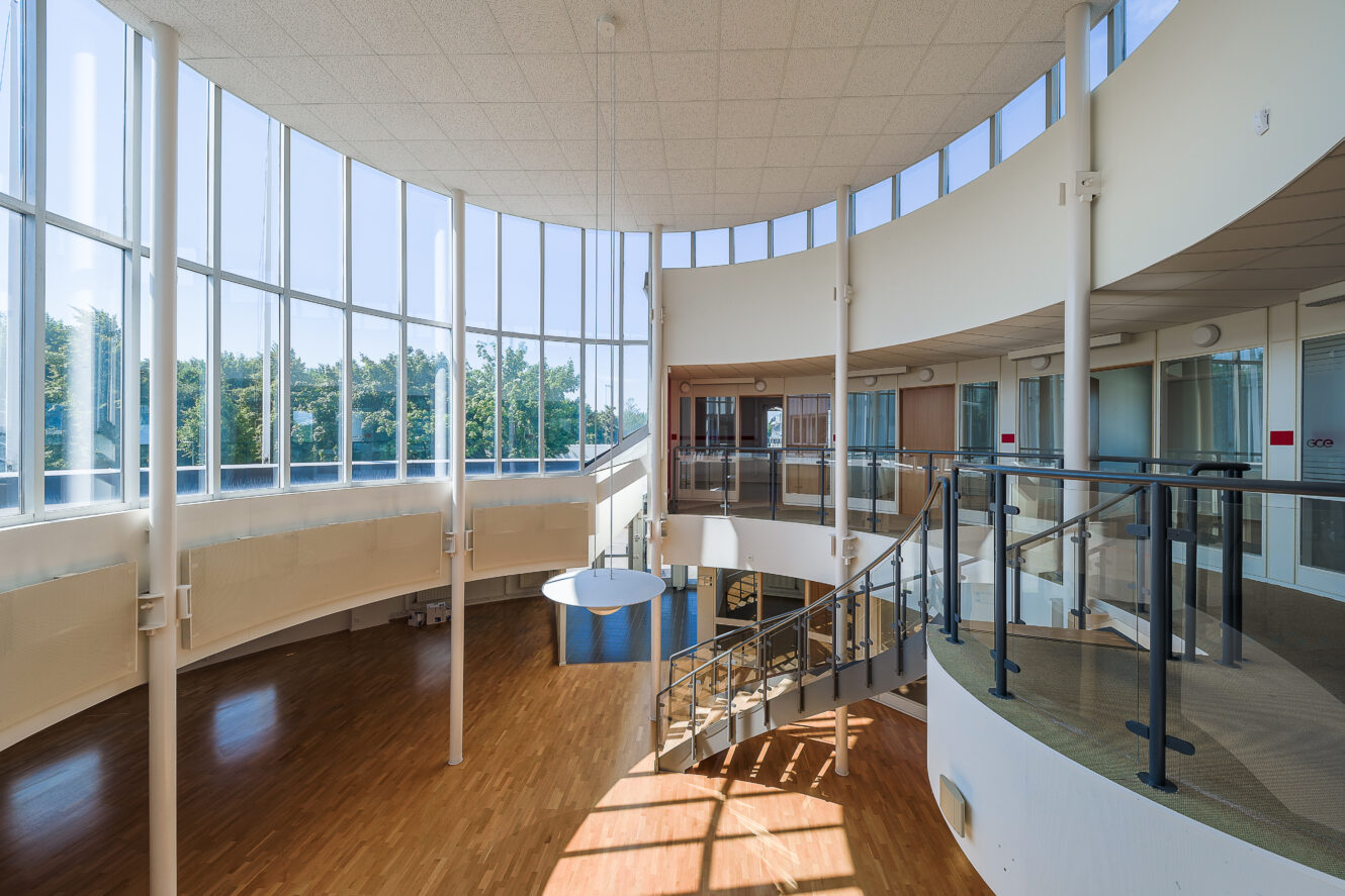 Rymlig, modern interiör med stora fönster från golv till tak, svängd trappa, glasräcken, trägolv och naturligt ljus som lyser upp utrymmet.