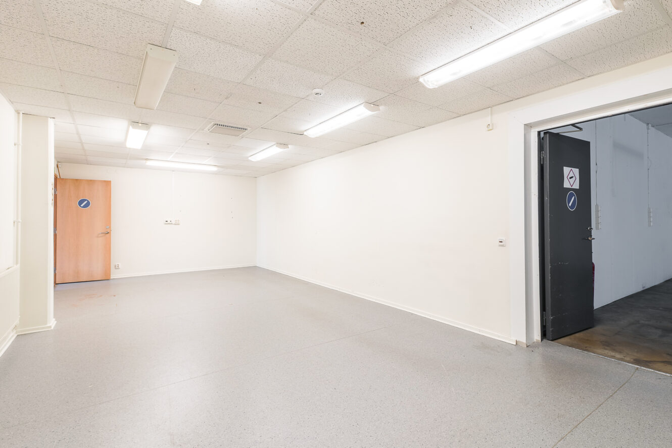 Tomt, väl upplyst rum med vita väggar, kaklat tak, grått golv, lysrör och två dörrar - en i trä och en i metall - som båda är delvis öppna.