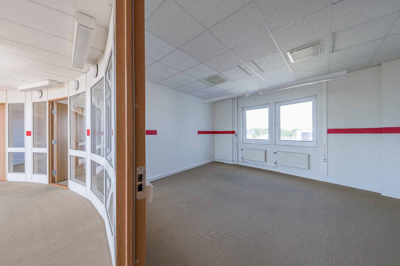 Ett tomt kontorsrum med beige matta, vita väggar, två fönster och en röd rand på väggen. En glasvägg med dörrar syns till vänster.