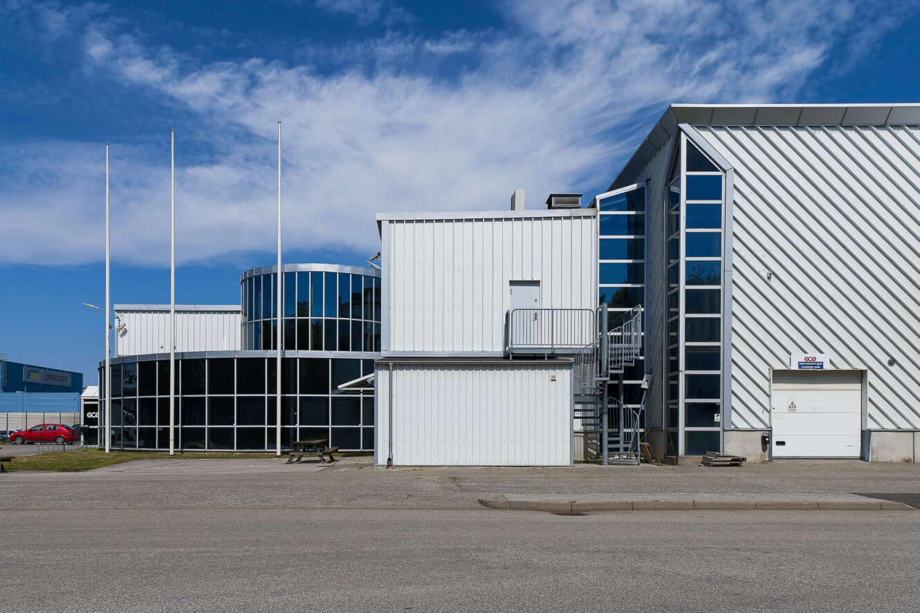 Modern industribyggnad med stora glasfönster, metallbeklädnad, en rund sektion, flaggstänger och en vit garageport under en blå himmel.