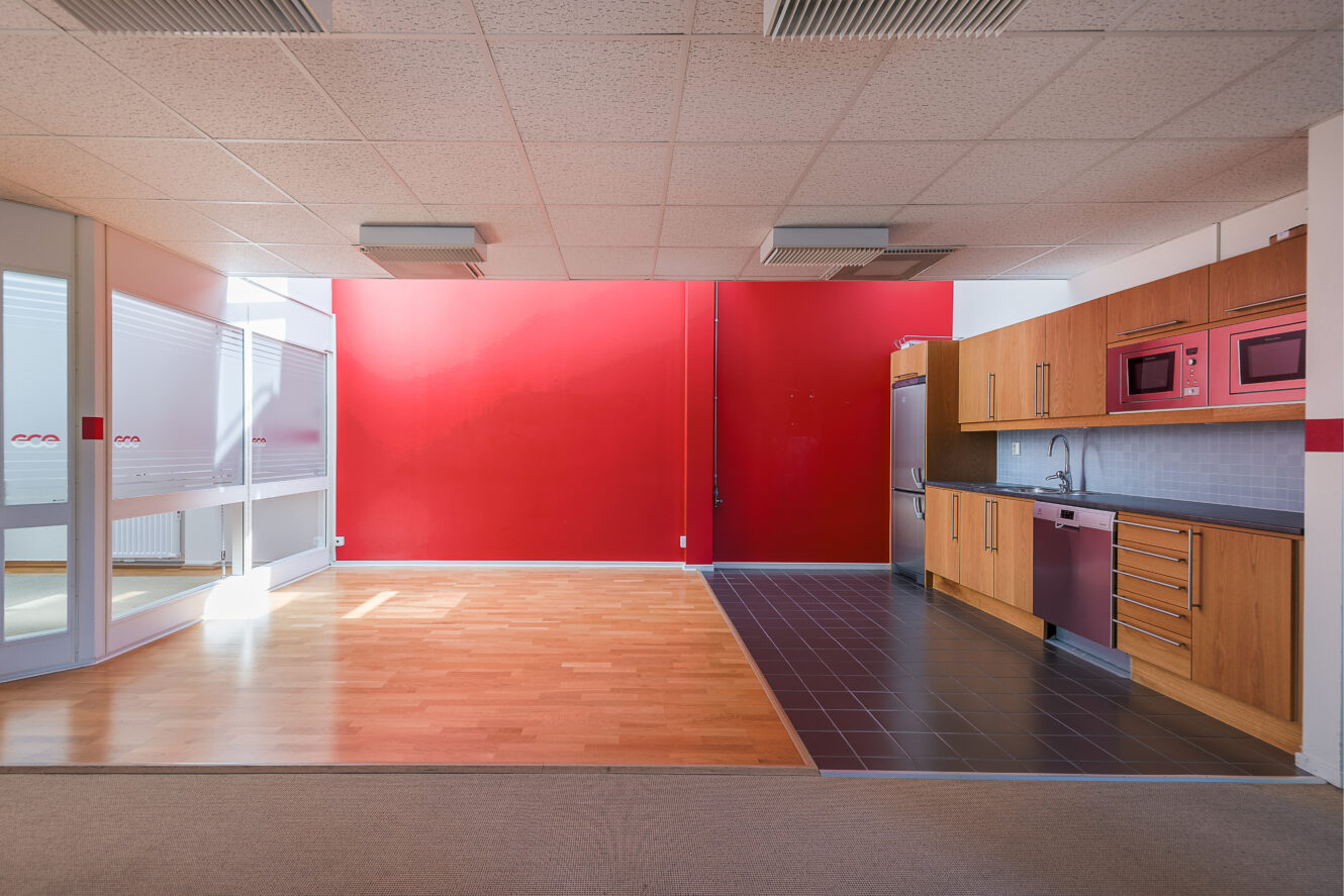 Modernt kontorskök med skåp i trä, vitvaror i rostfritt stål och en röd accentvägg, intill en öppen yta med trä- och klinkergolv.