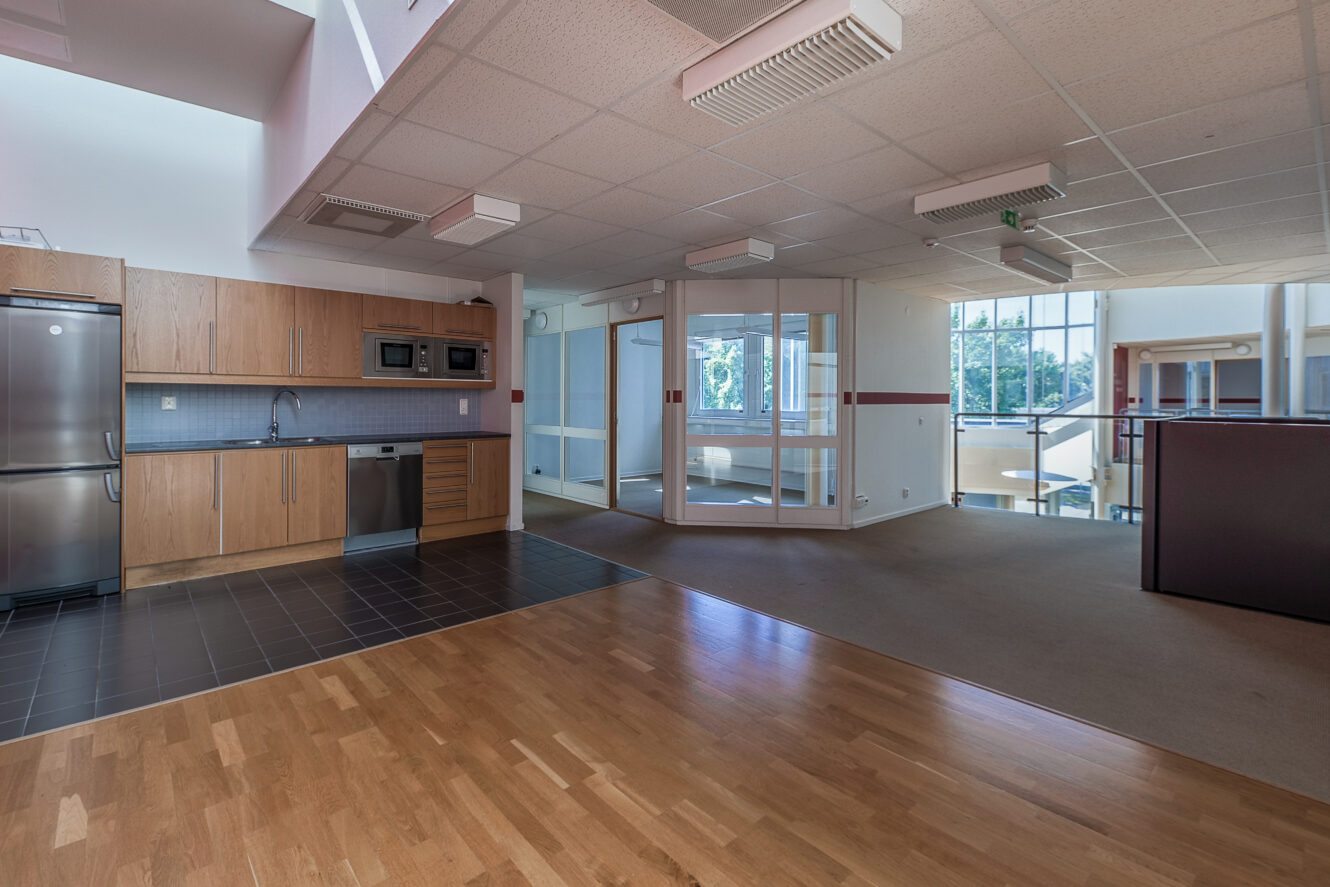 Modernt kontorsutrymme med ett litet kök, trä- och klinkergolv, glasväggar och naturligt ljus från stora fönster.