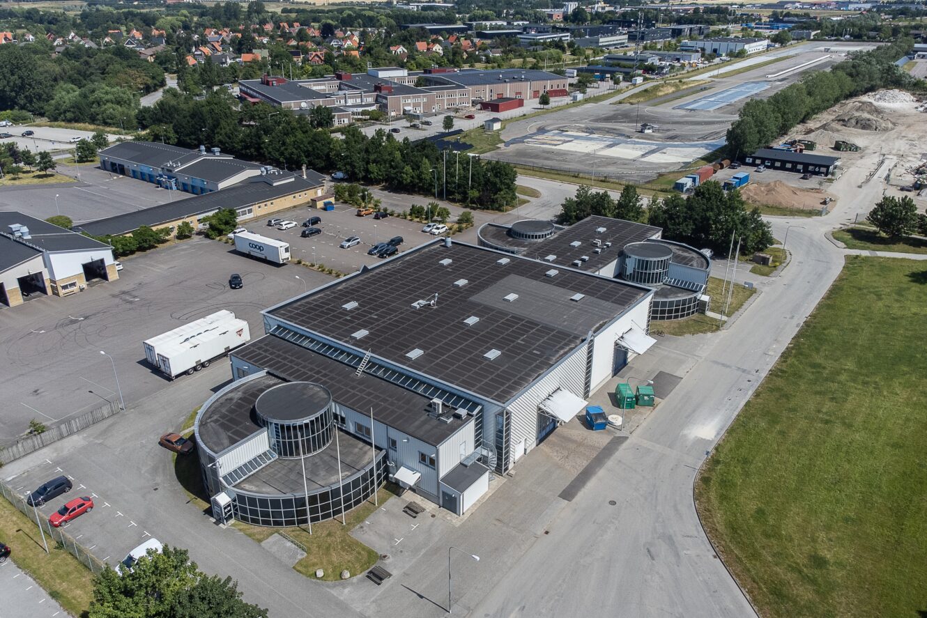 Flygfoto över en stor industribyggnad med svart tak och runda glasentréer, omgiven av vägar, parkeringsplatser och andra industrianläggningar.