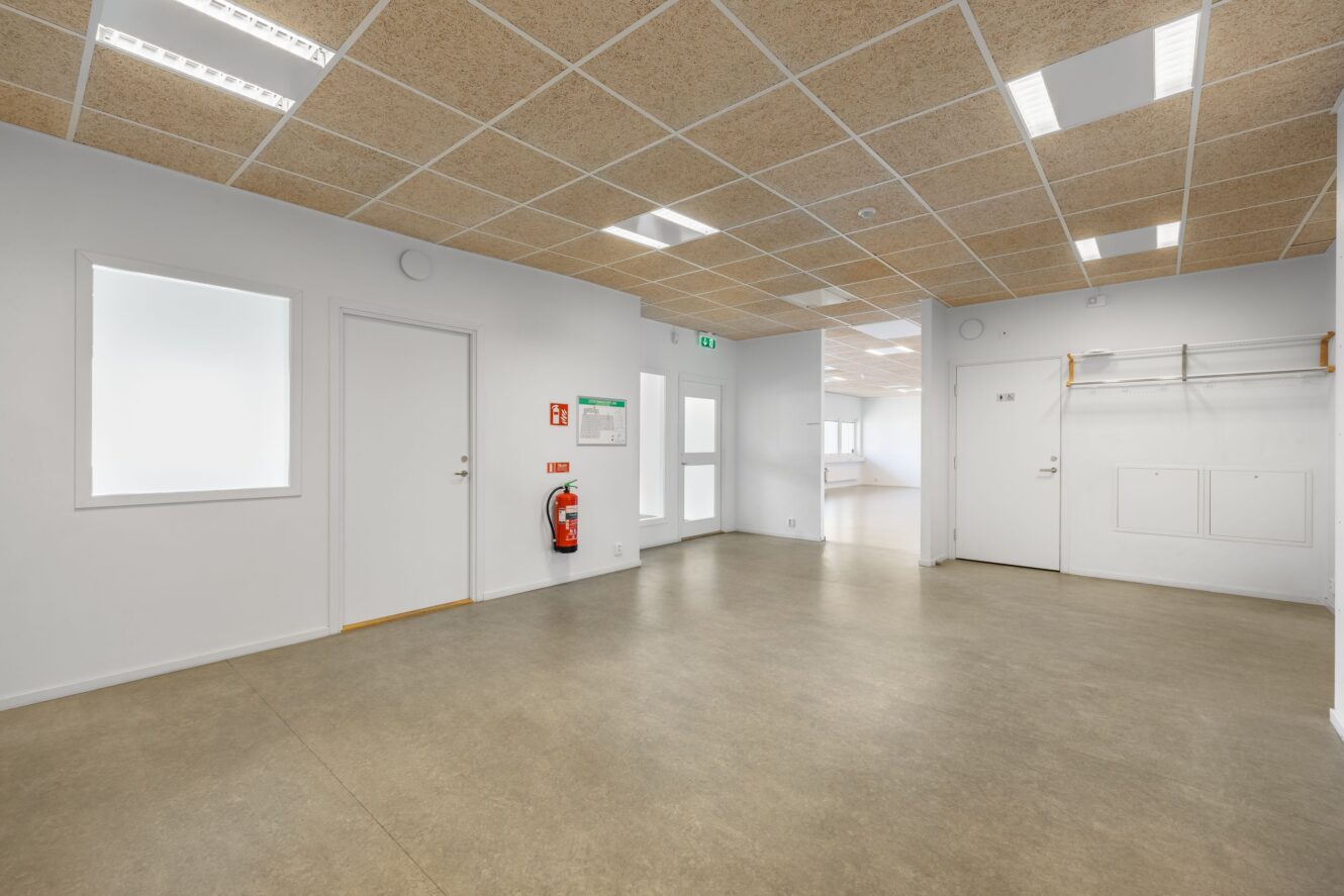 En ren, tom korridor i en modern byggnad med beige golv, vita väggar, en brandsläckare och lysrör i taket.