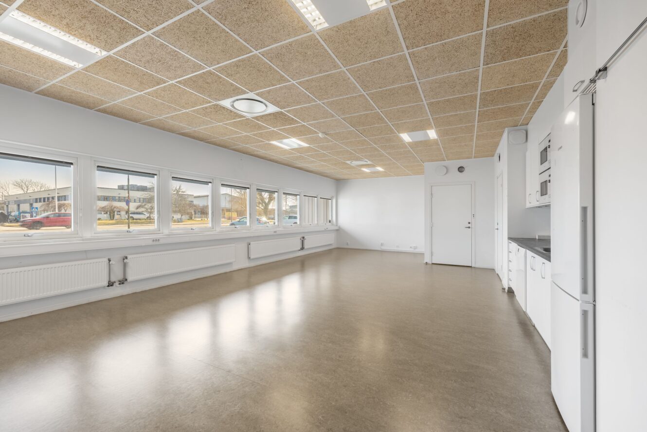 Rymligt, tomt rum med stora fönster, vita väggar, kaklat tak och ett pentry längs ena sidan. Utrymmet fylls av starkt naturligt ljus.