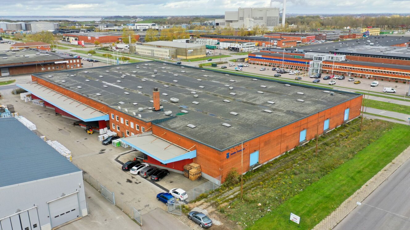 Flygfoto över ett stort industriellt lagerhus i rött tegel med lastkajer, parkerade fordon och omgivande industribyggnader.