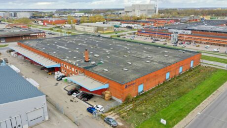 Flygfoto över ett stort industriellt lagerhus i rött tegel med lastkajer, parkerade fordon och omgivande industribyggnader.
