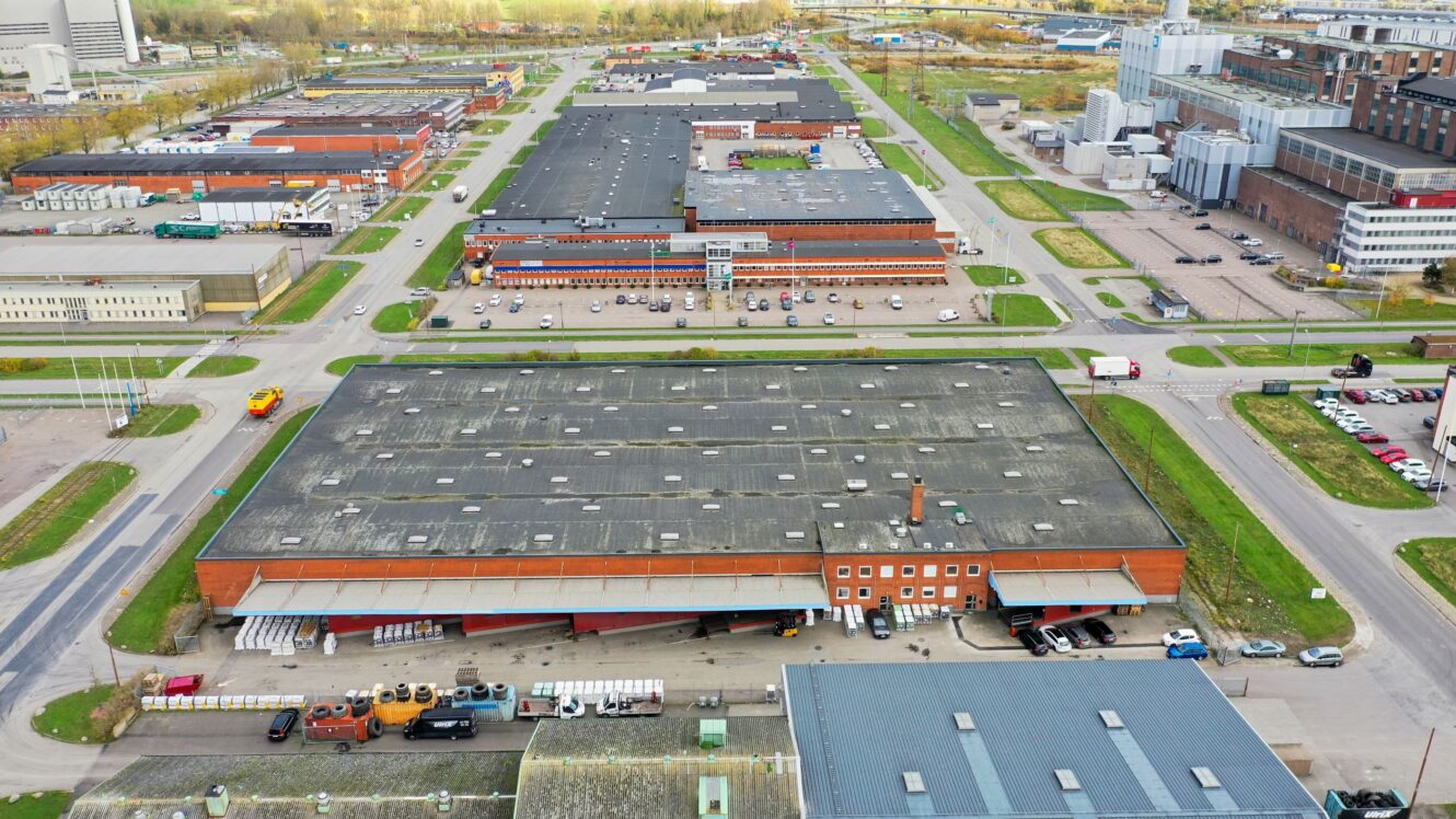 Flygfoto över ett industriområde med stora lagerbyggnader, parkeringsplatser, vägar och flera parkerade fordon en molnig dag.