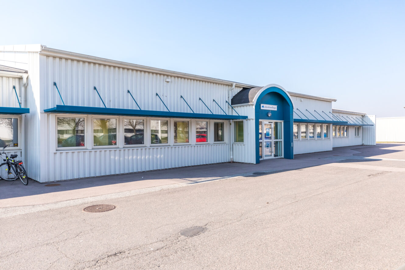 En envånings industribyggnad med vit metallfasad, blå dekor, en central välvd entré och flera fönster, sedd från parkeringsplatsen.