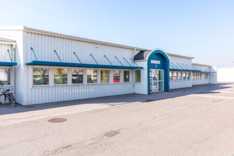 En envånings industribyggnad med vit metallfasad, blå dekor, en central välvd entré och flera fönster, sedd från parkeringsplatsen.