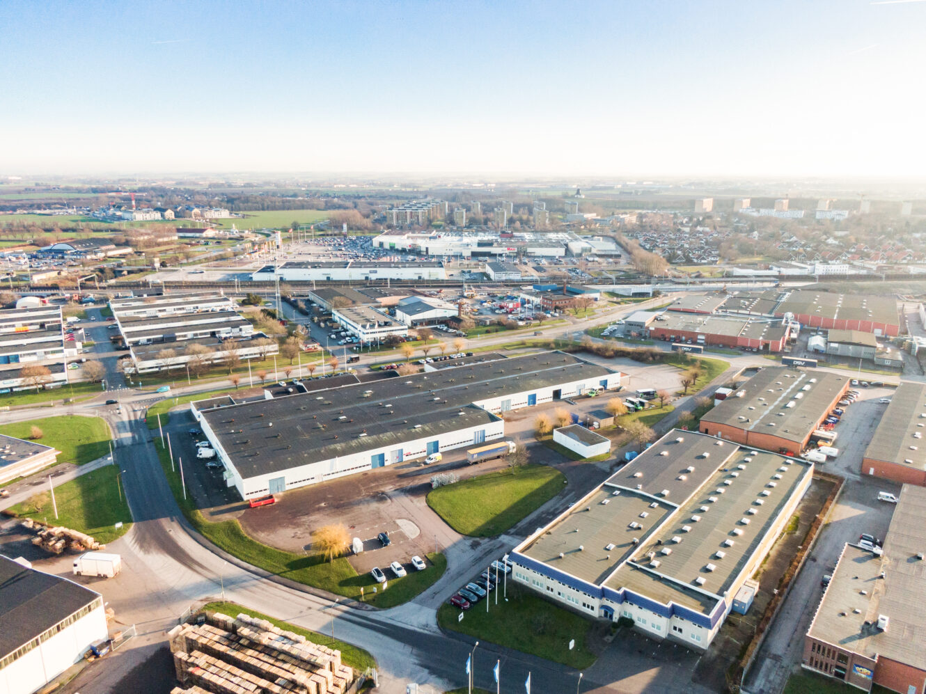 Flygfoto över en industripark med stora lagerbyggnader, parkeringsplatser, vägar och omgivande stadsområde en klar dag.