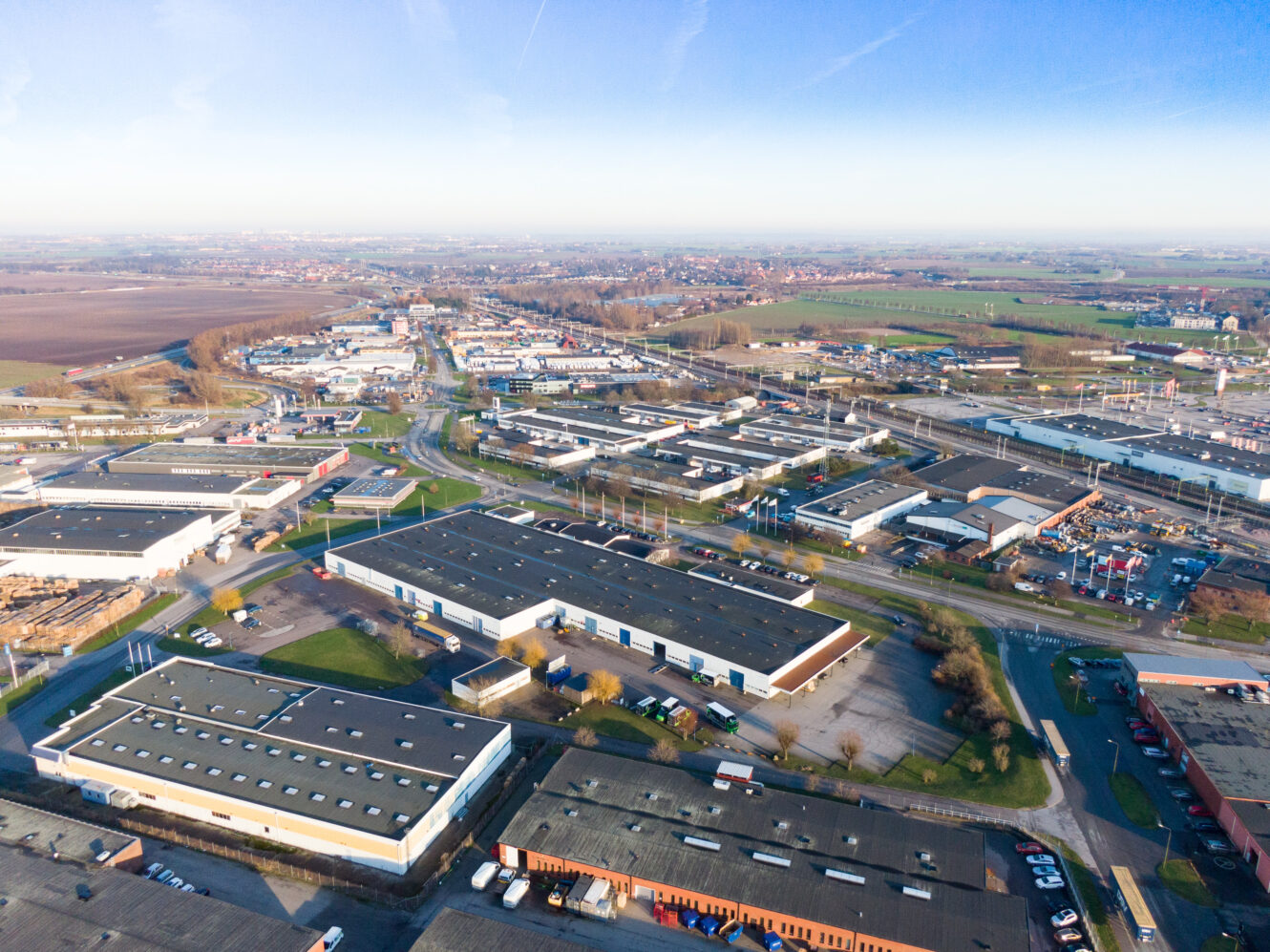 Flygfoto över en industripark med flera lagerlokaler, parkeringsplatser, vägar och omgivande fält under en klarblå himmel.