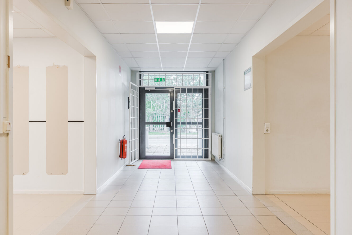 En ljus hall med vita väggar och klinkergolv, som leder till en utgångsdörr av glas med metallgaller. En röd matta ligger på golvet, och en brandsläckare är monterad på vänster vägg.