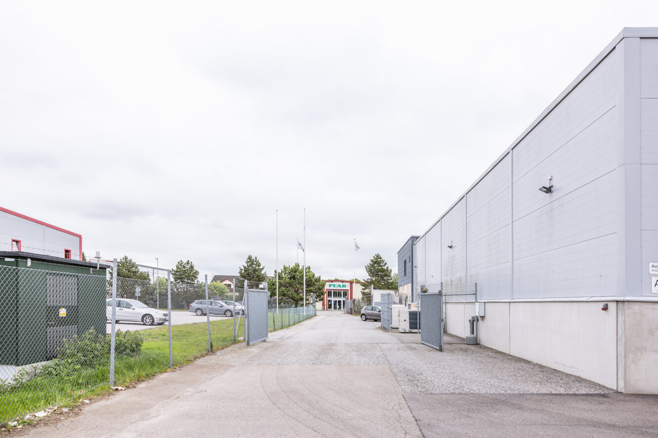 En asfalterad uppfart slingrar sig mellan två grå industribyggnader, med parkerade bilar och en inhägnad gräsyta till vänster under en molnig himmel.
