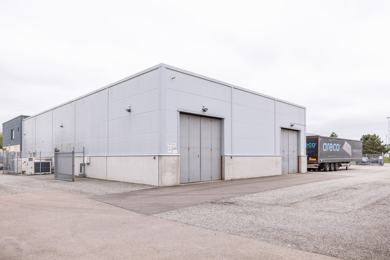 Ett stort grått industrilager med två stängda garageportar och en lastbil som står bakåtparkerad vid en av ingångarna; en tom asfalterad tomt framför, och en molnig himmel ovanför.
