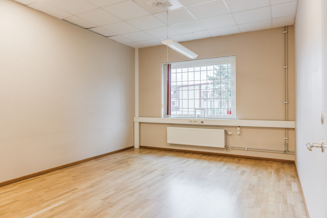 Ett tomt rum med beige väggar, ljust trägolv, ett fönster med galler, en radiator och en lysrörsarmatur i taket.
