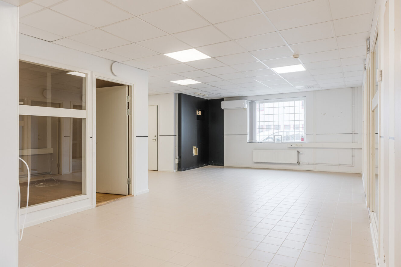 Ett tomt, rent rum med vita väggar, klinkergolv, ett stort fönster, en glasvägg, en öppen dörr och taklampor.