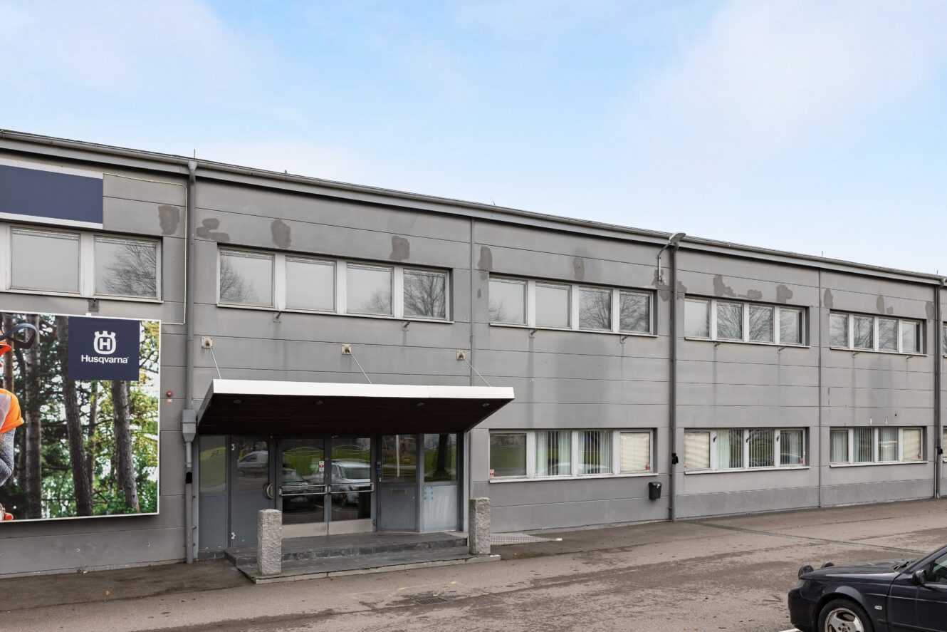 En grå affärsbyggnad i två våningar med stora fönster och en täckt entré, med en Husqvarna-reklam på vänster sida.