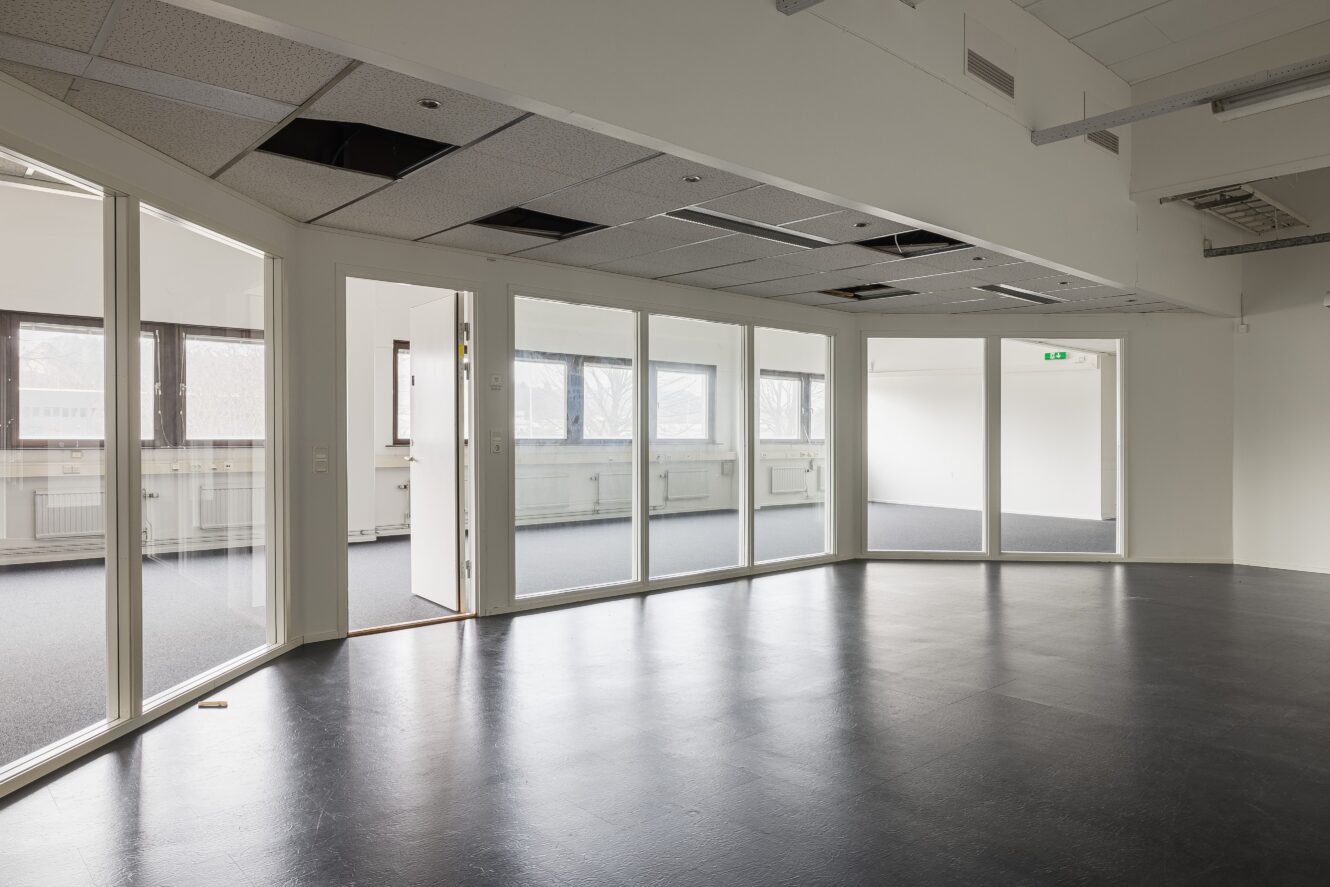 Tomt kontorsutrymme med glasväggar, takplattor och stora fönster som släpper in naturligt ljus i rummet.