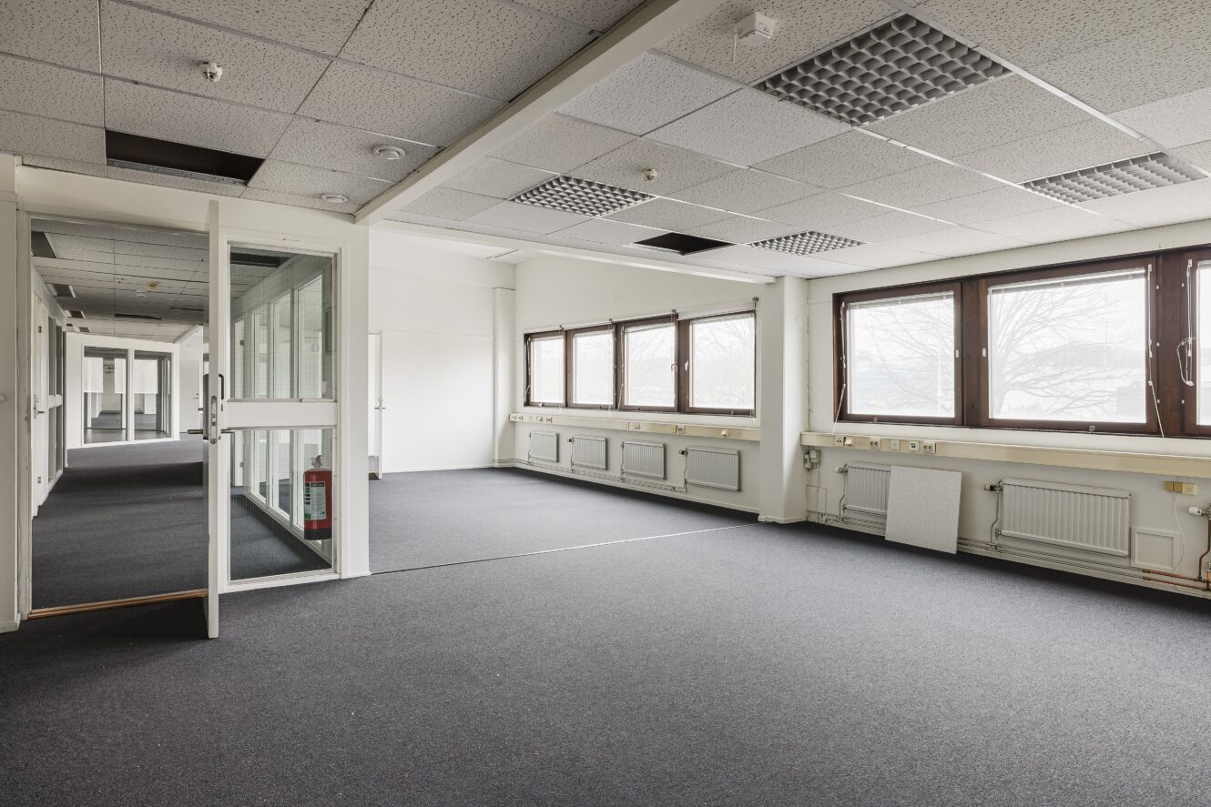 Tomt kontorsutrymme med stora fönster, grå heltäckningsmatta, vita väggar och en delvis öppen glasdörr; flera takplattor saknas eller har flyttats.