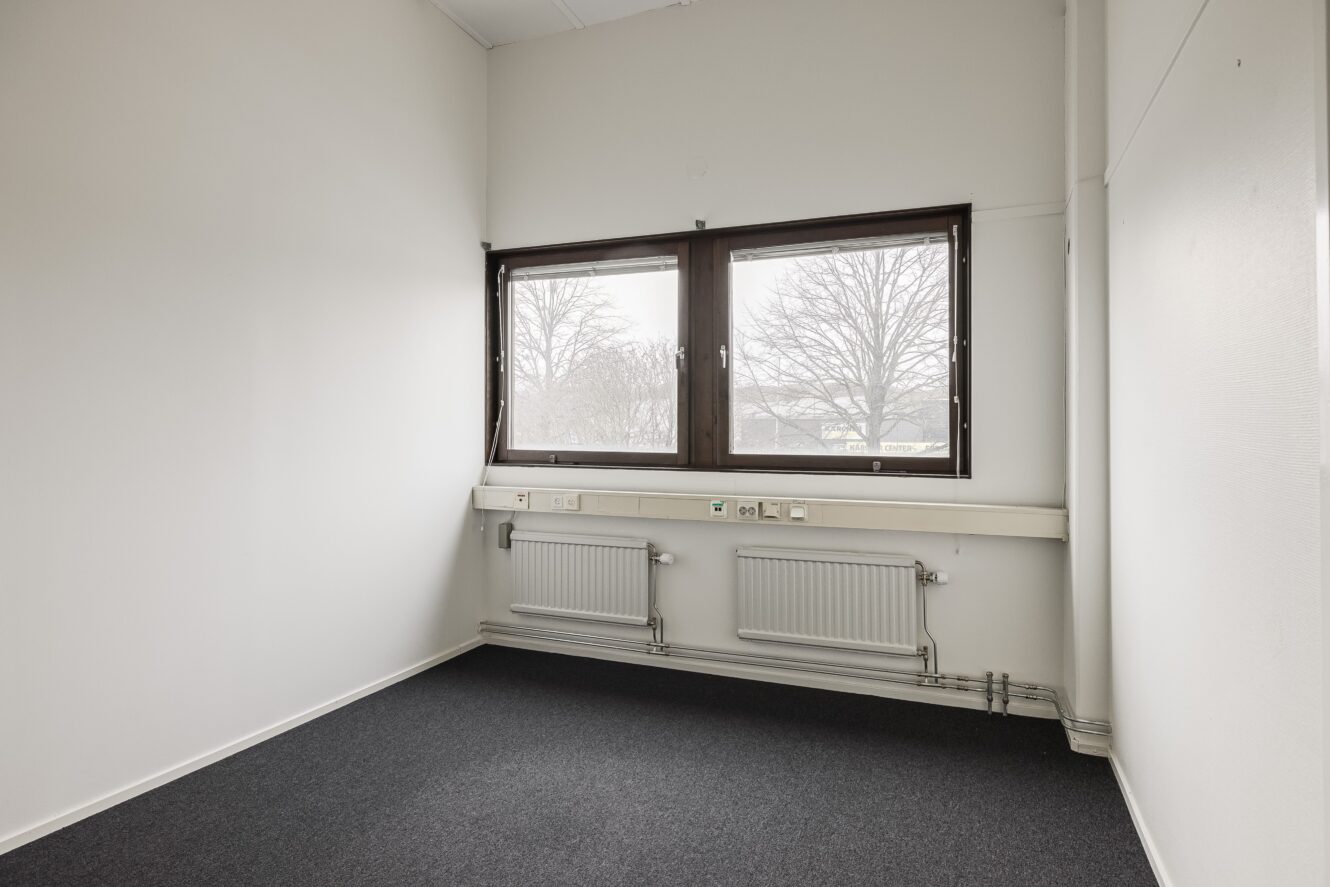 Litet, tomt kontorsrum med vita väggar, mörk heltäckningsmatta, två element, vägguttag och ett stort fönster med utsikt över kala träd utanför.
