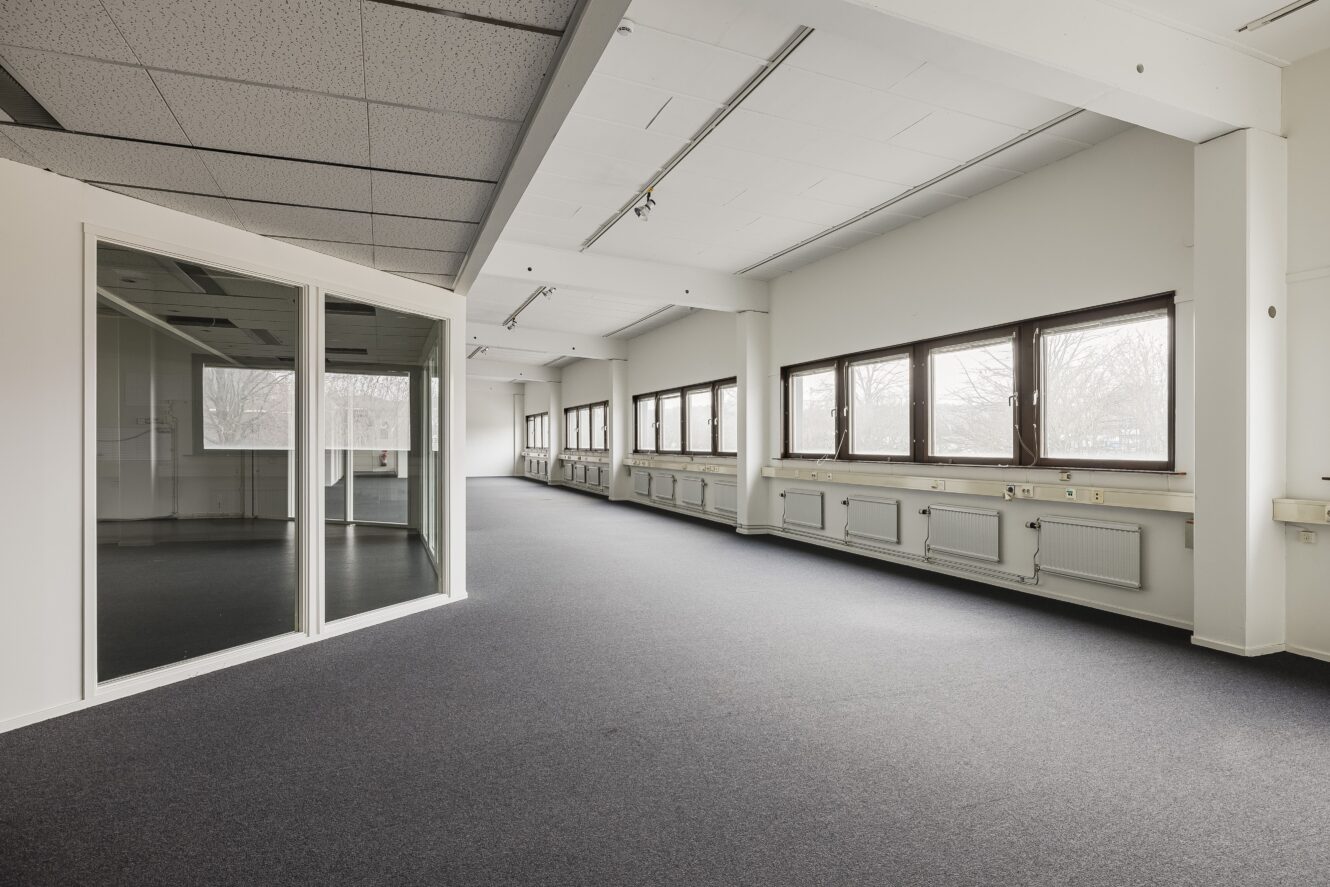 Stort tomt kontorsutrymme med grå matta, vita väggar, flera fönster och ett rum med glasväggar till vänster.