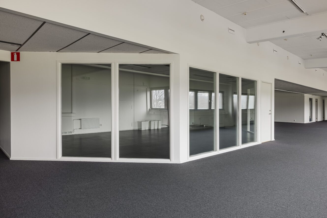 En tom kontorslokal med glasväggar, grå heltäckningsmatta, vita väggar och fönster som släpper in naturligt ljus.