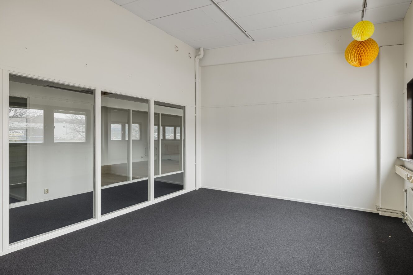 Ett tomt kontorsrum med vita väggar, mörk heltäckningsmatta, stora innerfönster och två gula papperslyktor som hänger i taket.