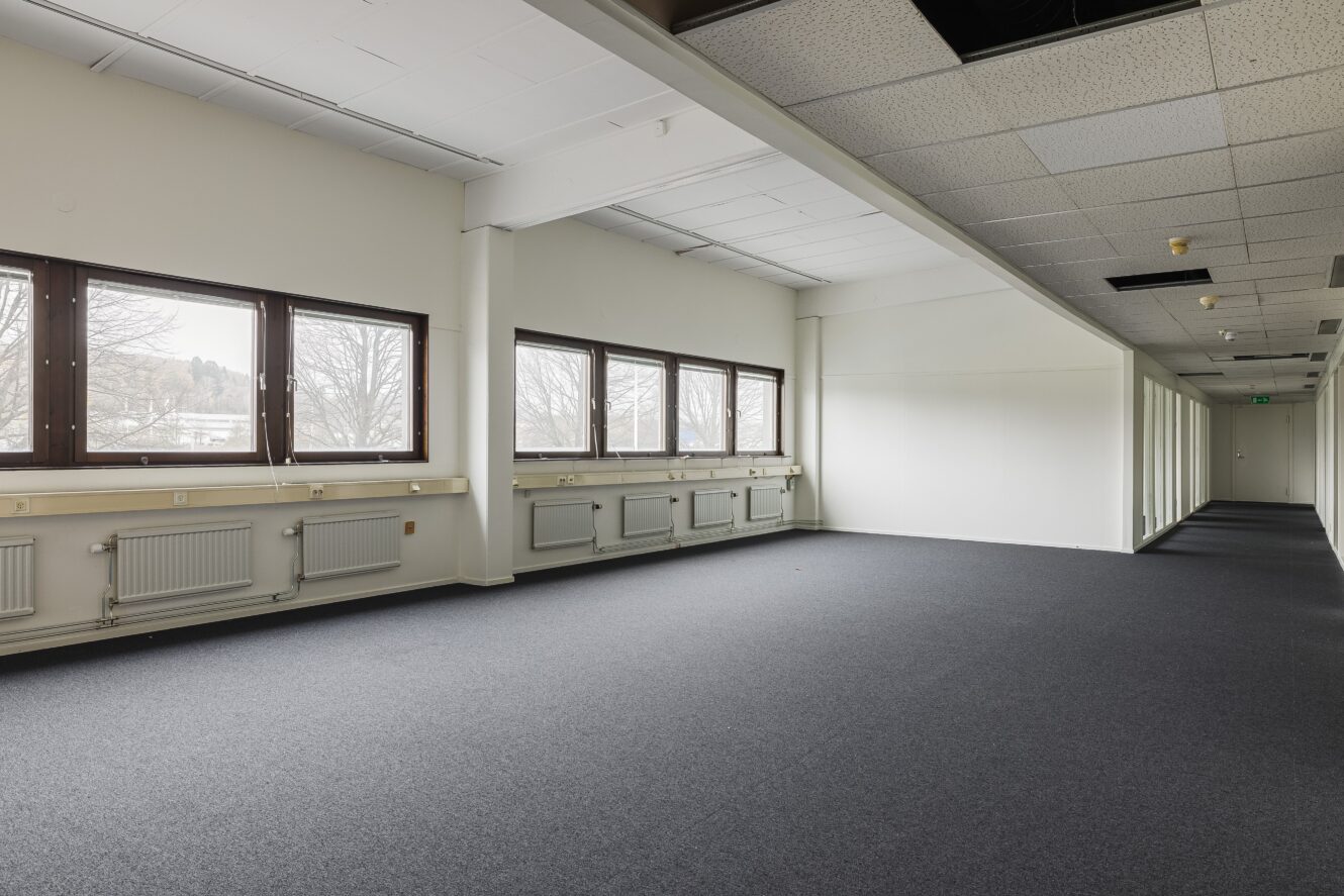 Tomt kontorsutrymme med grå heltäckningsmatta, vita väggar, flera stora fönster till vänster, radiatorer under fönstren och ett undertak där några paneler saknas.