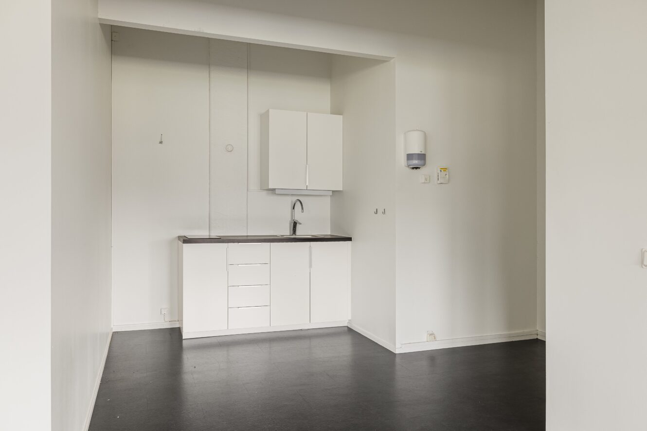 Litet, minimalistiskt pentry med vita skåp, diskho och en väggmonterad tvålpump mot vita väggar och ett mörkt golv.