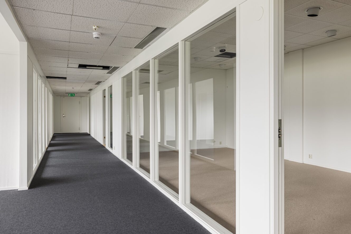 Tom modern kontorskorridor med glasväggar till höger, grå heltäckningsmatta och vita takplattor, varav några saknas eller är skadade.