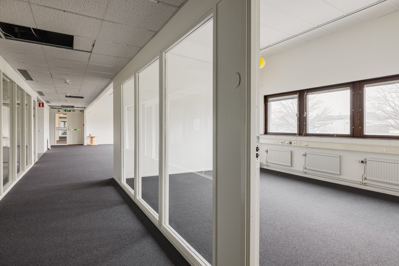 Tomma kontorslokaler med glasväggar, heltäckningsmattor, vita väggar och takpannor, stora fönster och radiatorer längs väggarna.