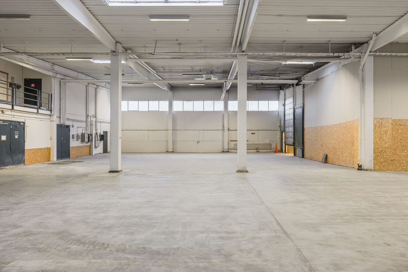 Tom lagerlokal med betonggolv, synliga bjälkar, vita väggar, högt i tak och lysrörsbelysning. Några elpaneler och en mezzanin syns på vänster sida.