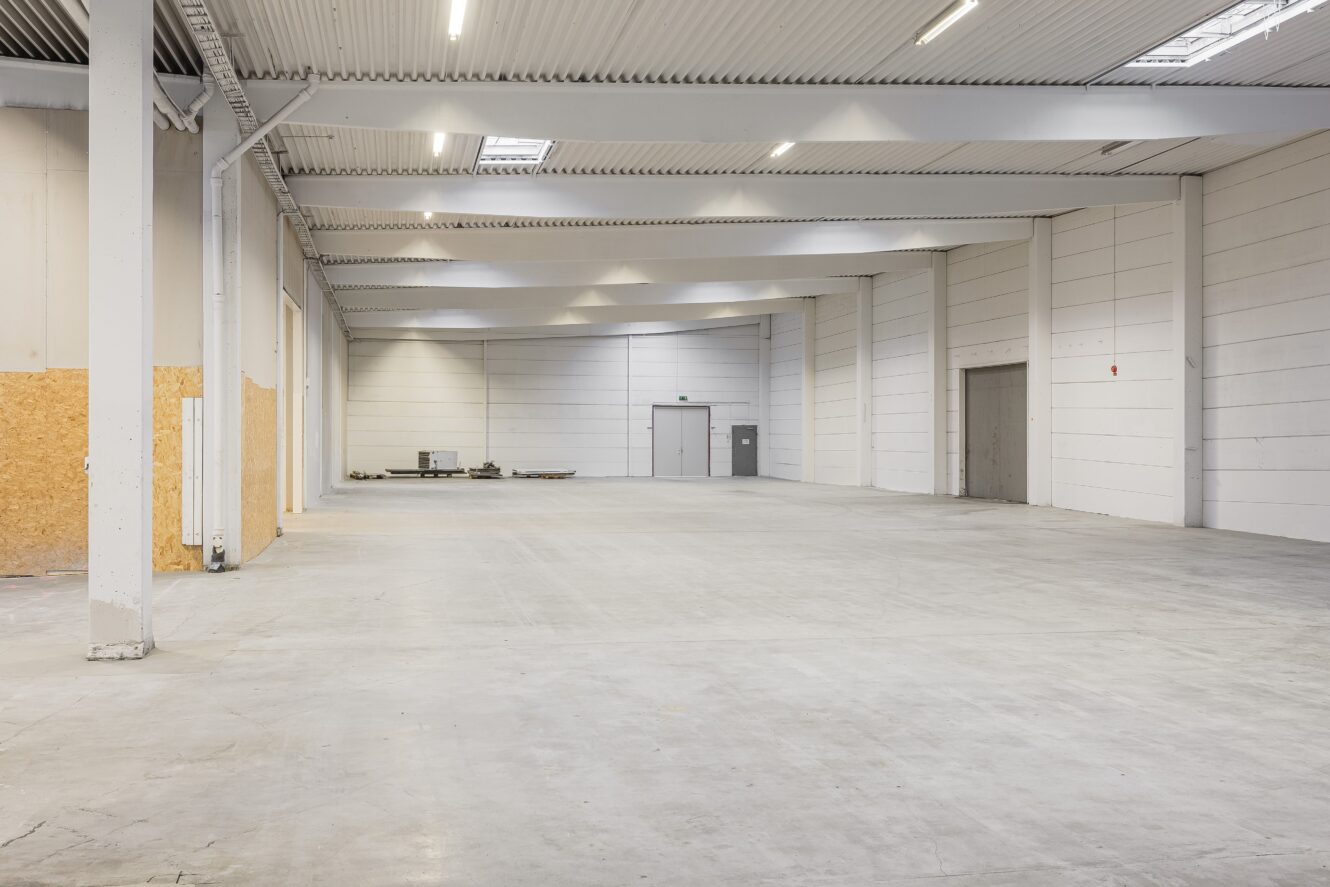 Stor tom lagerlokal med vita väggar, högt i tak, betonggolv, takbelysning och några föremål staplade i det bortre hörnet nära en stängd dörr.