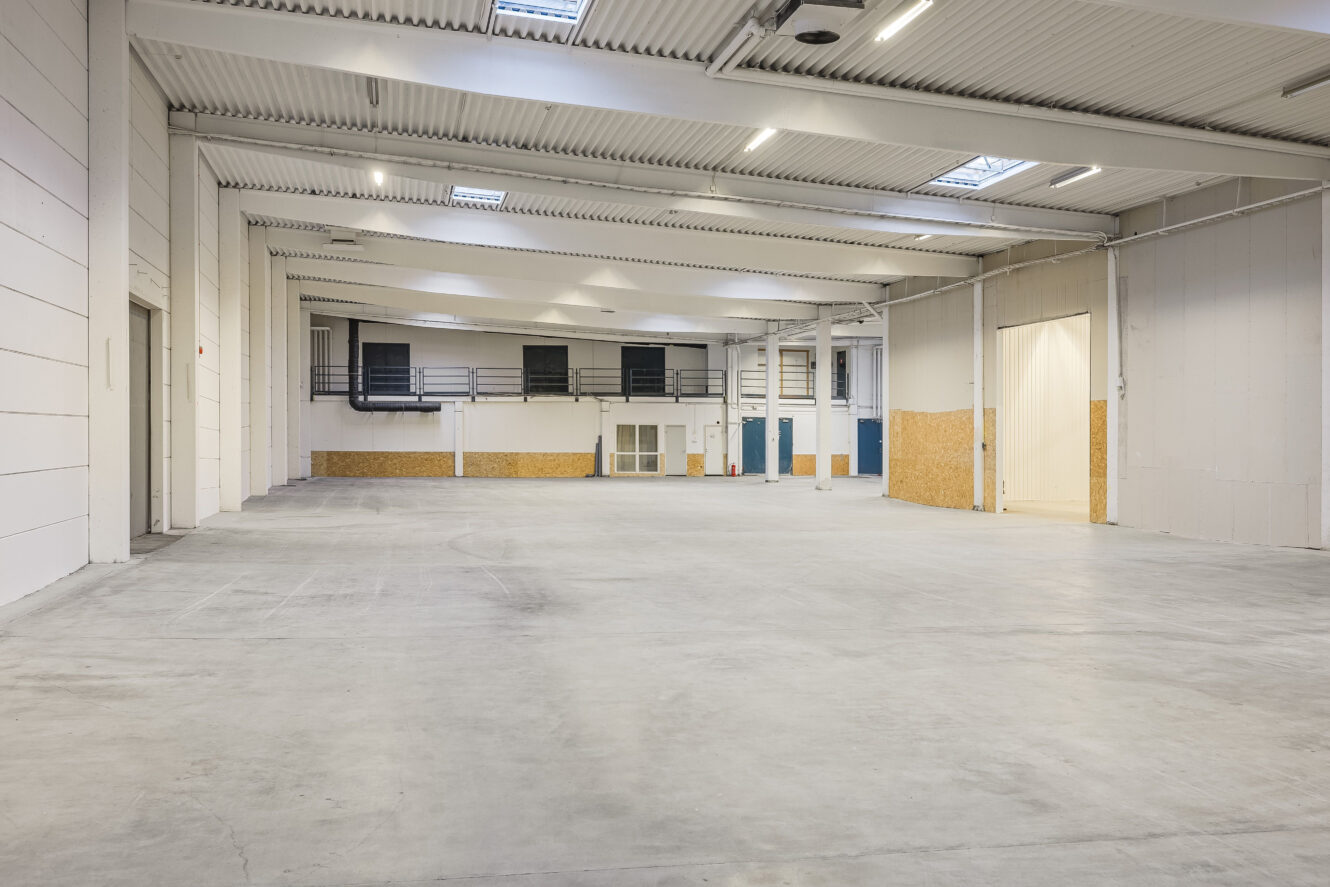 Stort tomt industrilager med vita väggar, högt i tak, betonggolv och ett litet kontorsutrymme på baksidan.