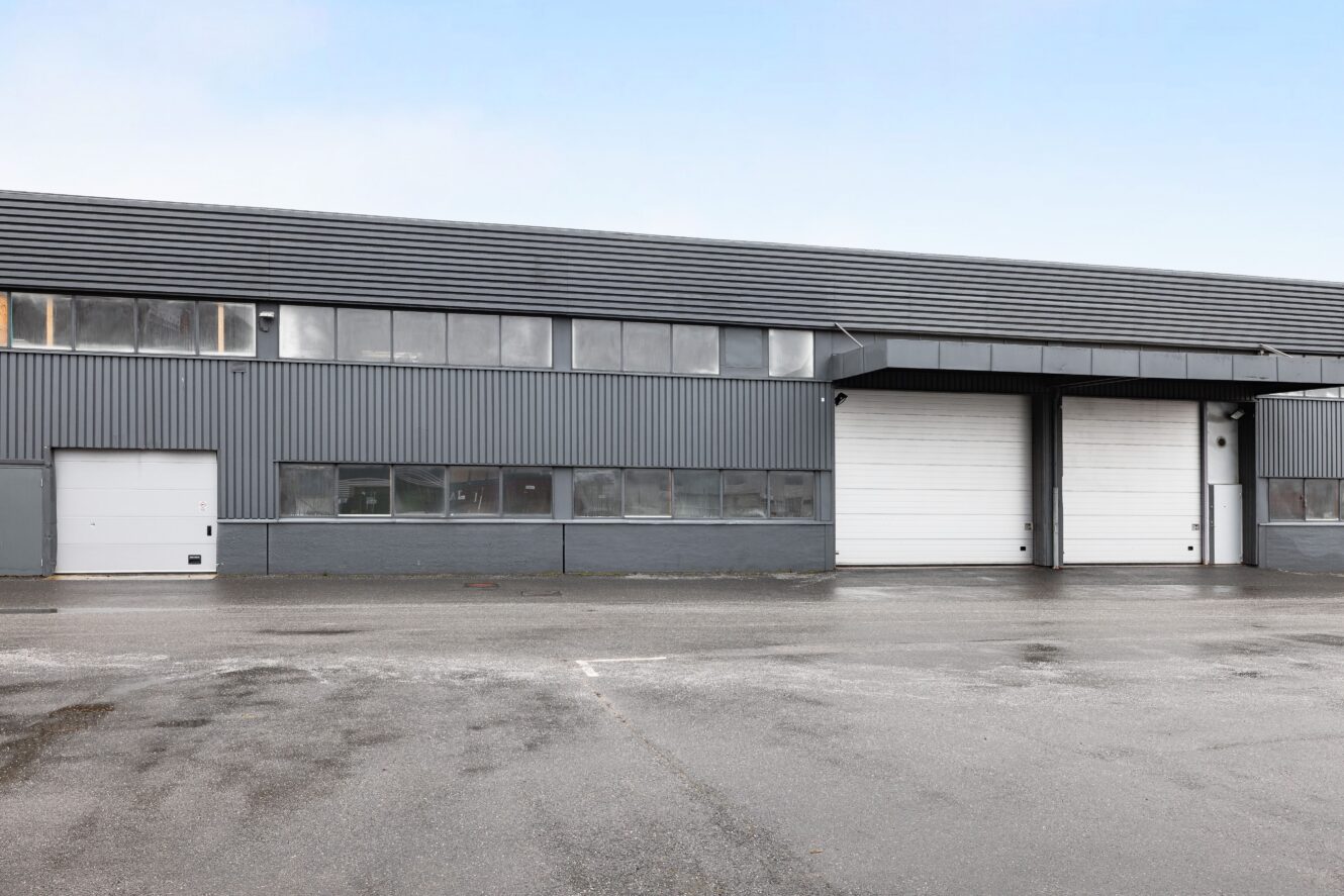 Grå industrilagerbyggnad med flera fönster, en liten entrédörr till vänster och två stora vita garageportar till höger; våt trottoar framför.